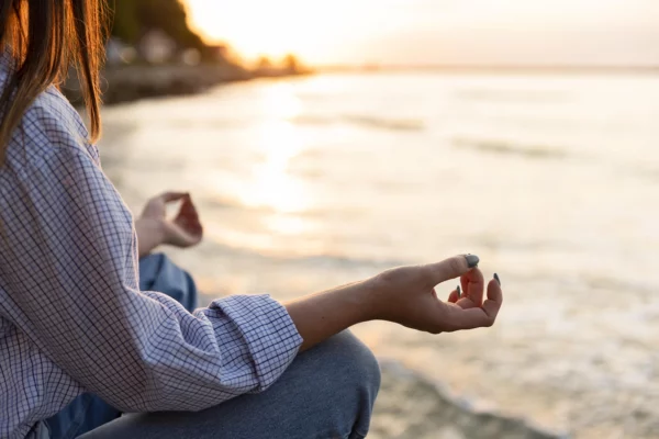 Femme méditant au bord de la mer au coucher du soleil, en position de relaxation avec les mains en mudra. Atmosphère paisible et harmonieuse, idéale pour la détente et la pleine conscience.
